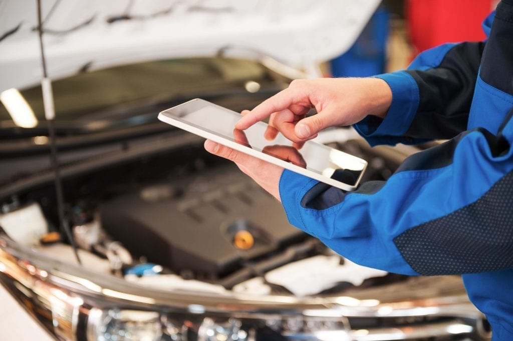 Close-up of man in uniform working on digital tablet while standing in front of car hood in vehicle service center